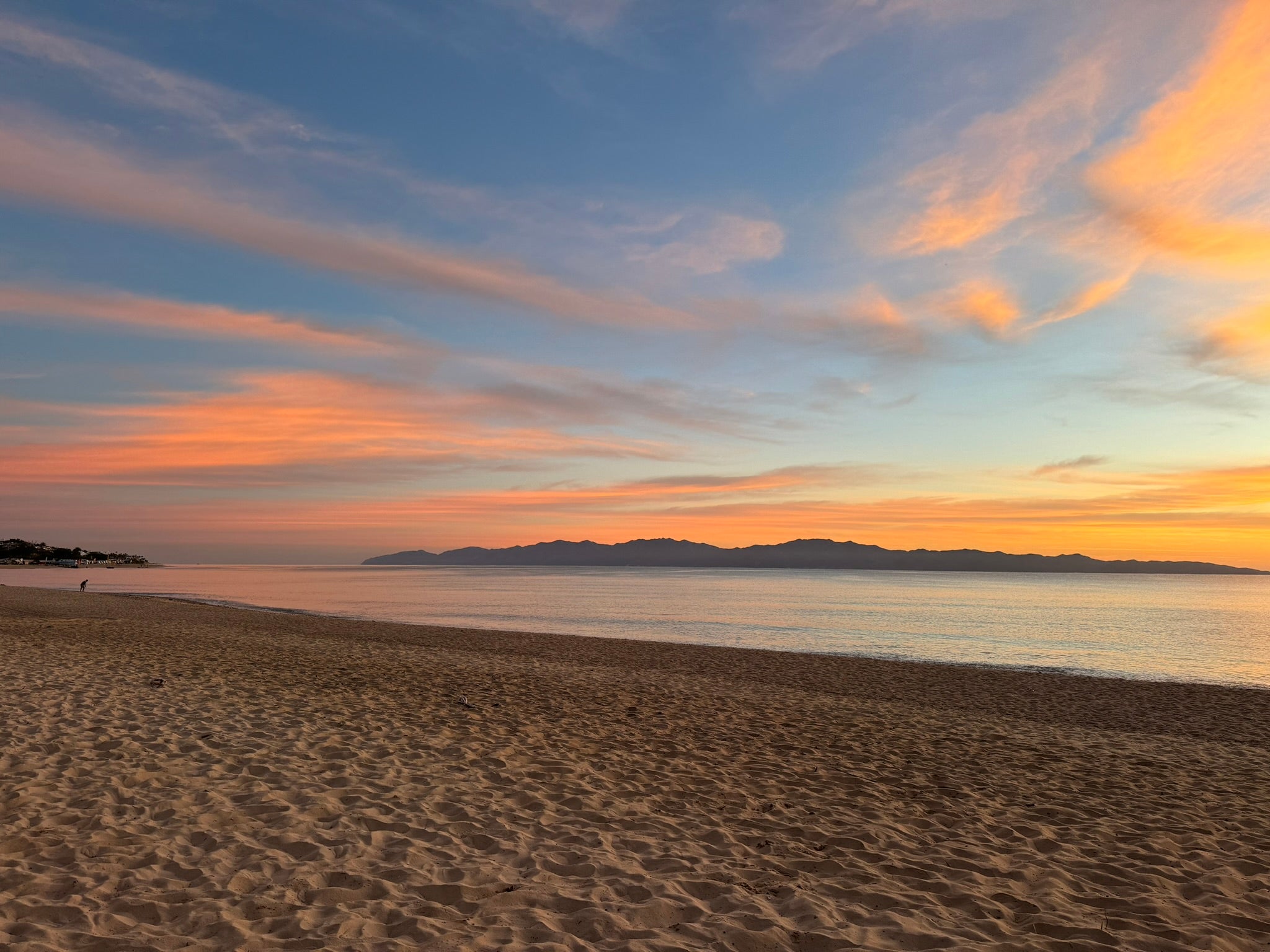 Peach and gold sunset clouds over a quiet beach, with mountains across the water.